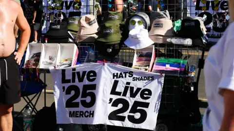 Emma Gibbs A bare-chested man stands next to a stall selling black, green and beige bucket hats and various sunglasses. It has a sign on it which reads: Live '25 Merch Hats Shades