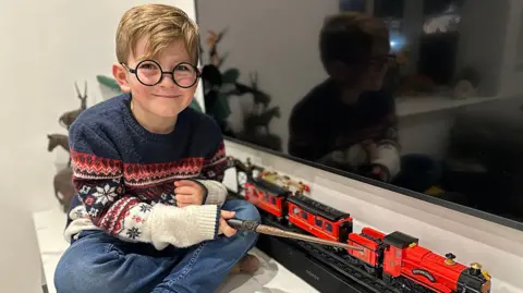 Richard, aged six, with blonde hair and blue eyes, holding his new wand, wearing 'Harry Potter'-style glasses, a festive Christmas jumper and jeans while sitting by a model of the Hogwarts Express at home