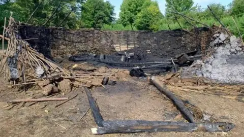 Flag Fen Wattle and daub walls of an Iron Age replica roundhouse with a pile of sticks and burnt wooden frame of an entrance and piles of ash and thatch.