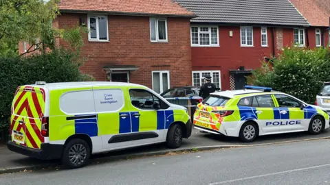 Michelle Adamson/BBC A crime scene investigation van and police vehicle outside a cordoned off house, which is being guarded by a police constable