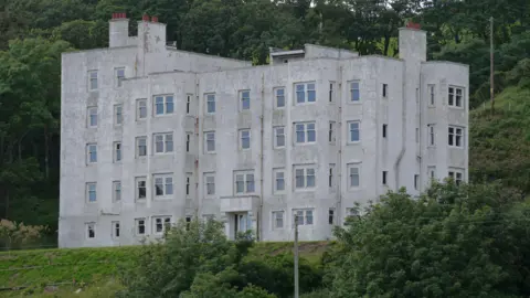 Geograph - James T M Towill A large five storey white building, once a hotel and now in a ruined state. It sits among trees and greenery.