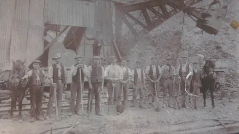 Forterra A sepia photograph thought to be from 1920 showing 11 men in white shirts and dark waistcoats stand looking at the camera. Two ponies are at either end of the line. All the men are holding spades, large sticks or the reins of the ponies. They are standing in front of a large machine looking directly at the camera.