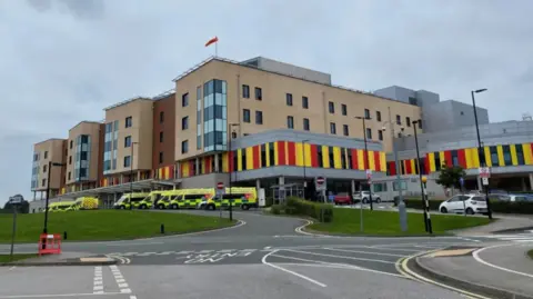 A large multi-storey building with multi-coloured cladding around the front. There are a number of ambulances outside