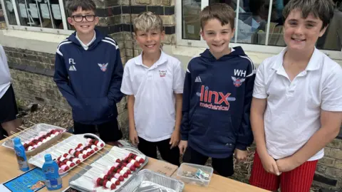 East Riding of Yorkshire Council Four schoolchildren standing behind a table, two wearing blue tracksuit tops and two wearing white polo shirts. There are a number of trays on the table containing what appears to be marshmallow kebabs.