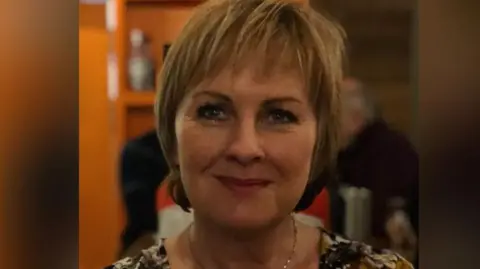 University of Worcester A woman with dark blonde shortish hair smiles at the camera. She is wearing a dress or top which is white with a flowery pattern and a necklace.