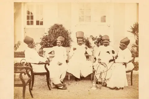DAG Five  men of India's Parsee community, dressed in traditional attire and distinctive headgear, sit on chairs in a garden, with a colonial-style building rising behind them.
