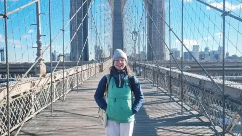 Becky Burke standing on the Brooklyn bridge, with a beige bobble hat and green body warmer, she looks at the camera smiling