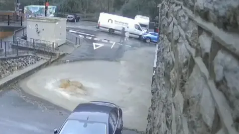 Two cars and a van close to a large pool of brown water covering a roadway next to a steep stone wall