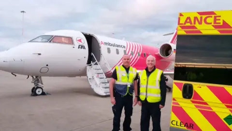 Lia's Wings Two ambulance men, wearing bright green jackets, smile in front of the medical plane