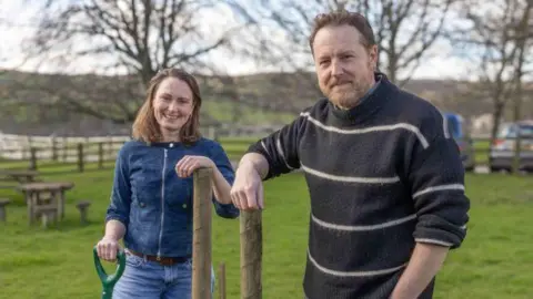 Yorkshire Dales National Park Authority Two people standing on a grassy field beside two newly placed wooden posts, one holding a green‑handled shovel, with picnic tables, bare trees and distant hills in the background.