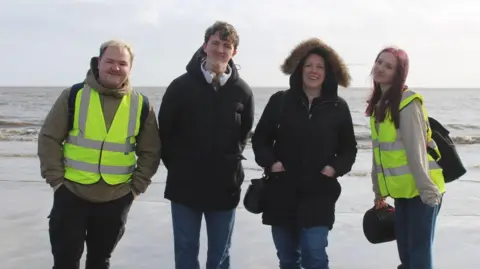 Madeline Jeffs Two men and two women are wearing coats, some also high visibility vests, and are standing in front of the sea on a beach.