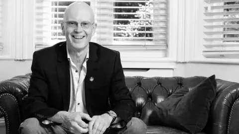 A black and white image of Rob Gittins sitting on a leather Chesterfield sofa at home. He is wearing a white shirt with a black blazer jacket and jeans. He is smiling and looking at the camera. 