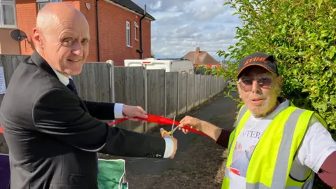 James Grant/BBC Mike Davey in a black suit is cutting a red ribbon while Chris Antoniou in a white T-shirt holds the ribbon