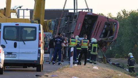 A crane winches an overturned bus up on the side of the road as firefighters look on. 