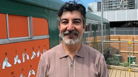 Hakan Elbir a smiling middle aged man with black hair and a greying beard stands in front of the cafe. There are residential blocks in the background. The the left is a coloured illustrations showing letters in sign language.
