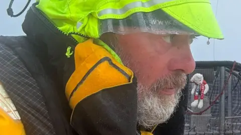 Bob Brown Bob has a grey beard and is looking away from the camera wearing waterproofs. 