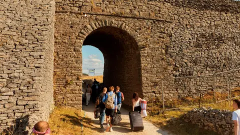 Joe Hart A stone archway with festival go-oers pulling suitcases and camping equipment through