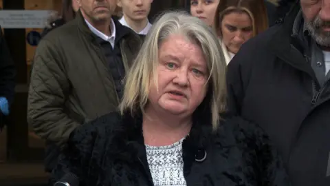 Wendy Cannon speaking outside the court. She has shoulder-length blonde hair and is wearing a black jacket and white and black patterned top. A number of people are standing behind her.