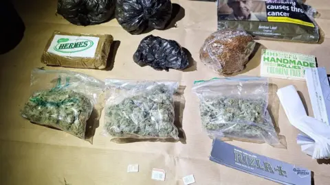 GMP Clear plastic snap bags filled with cannabis are lined up on a table. Black bin bag-style wraps are also seen displayed, alongside cigarette rolling papers, a pouch of tobacco and three phone SIM cards. 