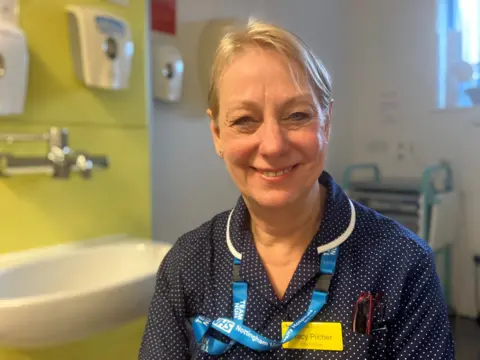 Tracy is smiling at the camera. She has blonde hair tied back from her face and is wearing a navy uniform with small white dots. Around her neck is a blue NHS lanyard and she has a bright yellow name badge on her chest. The background is a blurred medical room with a sink against a yellow wall and a window just visible to the right of Tracy.