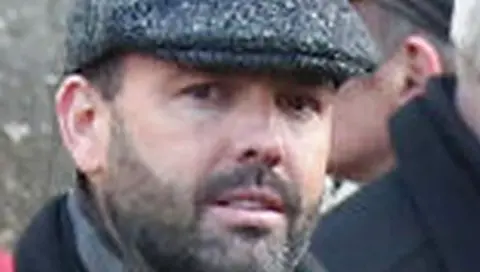 Man looks at camera, he has dark stubble and is wearing a flat cap