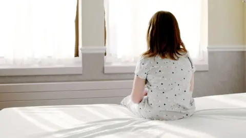 A girl wearing polka dot pyjamas, sat on the edge of a bed with a white duvet. She is looking out towards two windows with white curtains. 