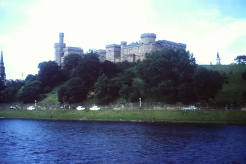 Ray Stokes Castle  behind trees in front of a river, with old cars on a road in between.