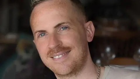Steven Elliott is smiling at the camera. He has a moustache and a light beard and is wearing a thick chain around his neck