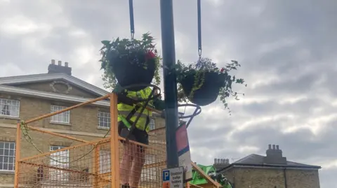 Mousumi Bakshi/BBC Hanging baskets being placed on street lamps by a person in a high-visibility jacket