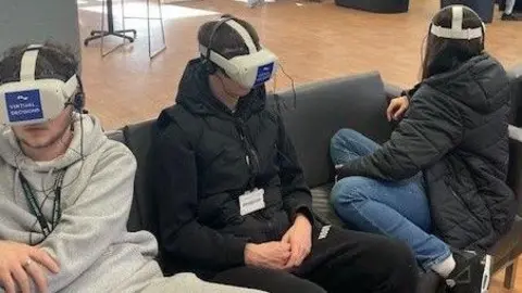 Hertfordshire Constabulary Three pupils from Oaklands College, sitting on a sofa, with VR headsets on. One is wearing a grey hoodie, one in the middle is wearing all black and the person on the left is facing away from the camera and is wearing blue jeans and a black coat. 