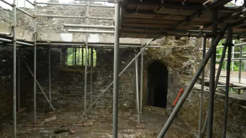 Inside Betsy Grimbal's Tower, metal scaffolding surrounds weathered stone walls and a small arched doorway, giving a view of the medieval structure during restoration work.