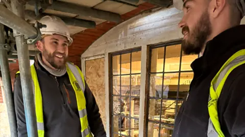 Tom and George Bayliss, looking at each other and smiling. They both have beards, and are wearing white hard hats, black tops and high-vis jackets. They are standing by a window, under scaffolding. 