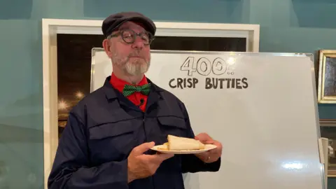 A man in a blue lab coat holds a crisp butty next to a sign reading "400 crisp butties".
