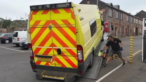 Amy Holmes/BBC Yellow emergency response vehicles with rear chevron markings being loaded onto a recovery truck by a worker in an urban industrial area with boarded-up brick buildings in the background.