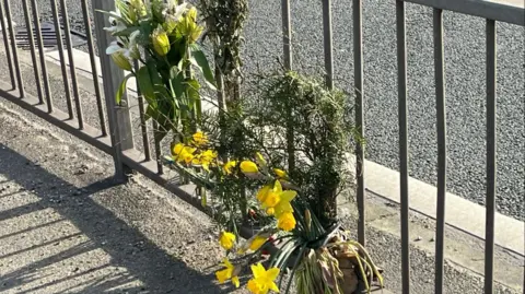 A selection of yellow and green flowers tied to a railing next to a road.