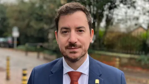 Sam Read/BBC Mike Reader standing outside looking directly to the camera and smiling. He is wearing a blue suit jacket, light coloured shirt and red tie.