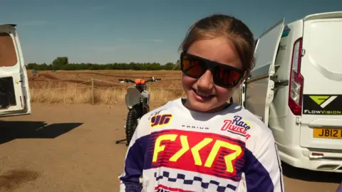 A young girl wearing sunglasses and a long-sleeved sports top with a variety of logos stands in front of a motocross bike and two white vans.