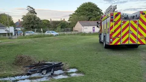 Withernsea AFC A fire engine parked on a playing field, facing away from the camera. On the left is the remnants of a small fire - burnt wood and sticks - on green grass.