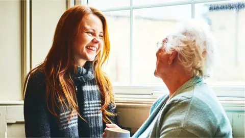 Getty Images A granddaughter and her grandmother smile together beside a window