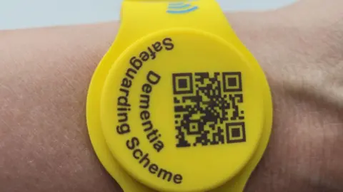 Cambridgeshire Police A close-up of a yellow watch-like wristband on someone's arm. The circular watch-face part has black writing on it that reads "Dementia Safeguarding Scheme" and it also has a QR scanning code on it.