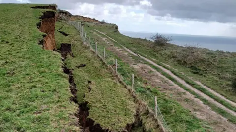 The coast path has a wood post and wire fence running along it and there is a major landslip leaving deep trenches in the grass and the soil exposed. The sea is beyond.