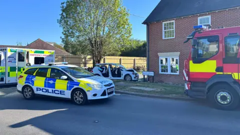 Matt Marvel/BBC An ambulance, a police car, and a fire engine parked outside a house and at the scene of a crash involving a white car, which is located in the middle of the picture and is badly damaged. 