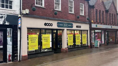 A cream coloured shop front with a red brick exterior above is pictured on a high street on a wet day. The store has a green sign that reads Robin Elt Shoes. 