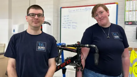 Tom Jackson/BBC Rob is standing on left and has short brown hair and is wearing black framed glasses. He is wearing a navy T-shirt which has a TAG BIKES logo on it. In the middle is a bike, and standing on the right of that is Lizzy, who is wearing the same T-shirt and has short blonde hair.