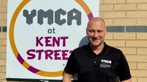Rob Baty stood in front of the "YMCA at Kent Street" sign, smiling at the camera and wearing a black T-shirt with "YMCA" written on in white letters.