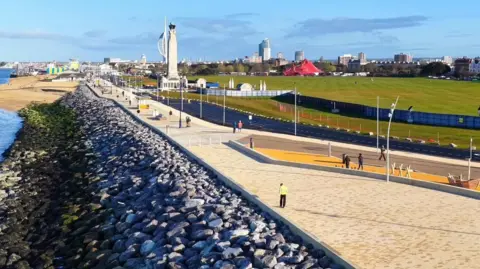 A wide seaside promenade separates a rock revetment on the shore and Southsea Common