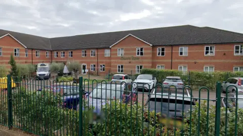 Google The outside of Olive Row, a care home in Kettering. The building is a brown brick residential building with a car park outside of it. The building backs onto a road and has a metal fence and bushes outside of the parameter. 