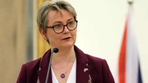 Yvette Cooper, who has cropped blonde hair and wears dark-framed glasses, speaks at a lectern in front of a British flag 