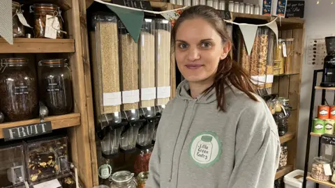 A woman is standing in front of containers that have dried foods in them. She has red-brown hair tied in a ponytail and is wearing a green hoodie. There is a green jar logo that says "little green pantry" on it.