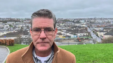 Shows a man with glasses and a brown jacket, white t-shirt, open-neck shirt and grey zip-up sweater standing in front of a grassy area. Behind, in the distance you can seee a digger on a derelict site and houses in the background.